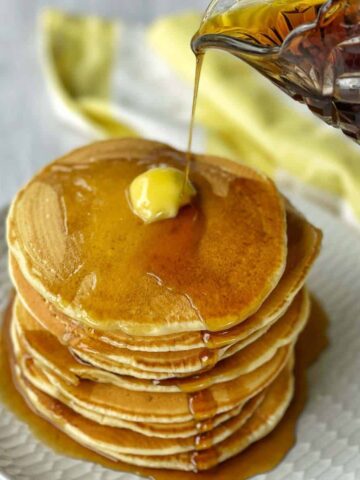 A stack of pancakes with butter on the top being poured with maple syrup.