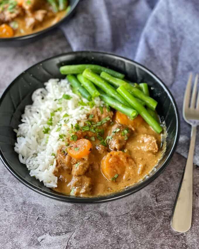 Slow cooked beef curry served in a black bowl with rice and green beans.