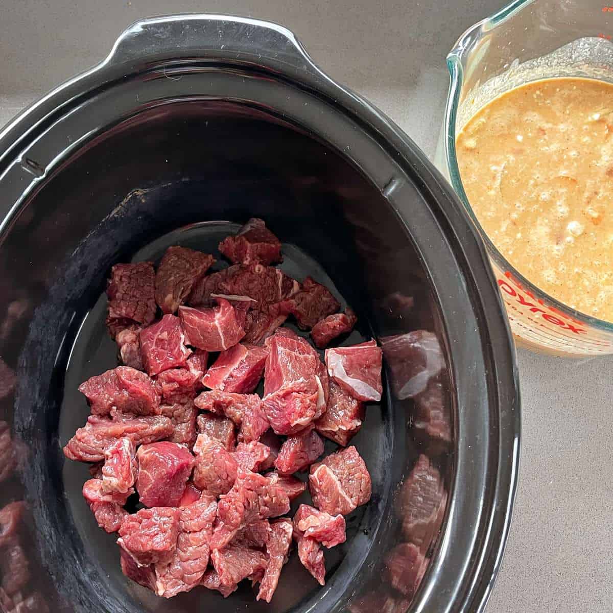 Chooped beef pieces inside a slow cooker. The curry mixture in a glass jug to the side.