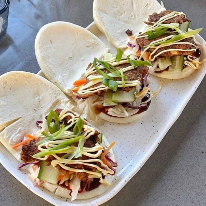 Three Crispy Chicken Bao Buns on a long white platter. The buns are open showing the chicken and salad filling.