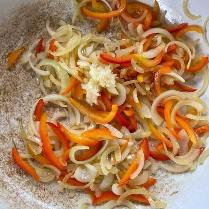 Sliced onion, sliced red pepper and grated garlic frying in a ceramic dish.