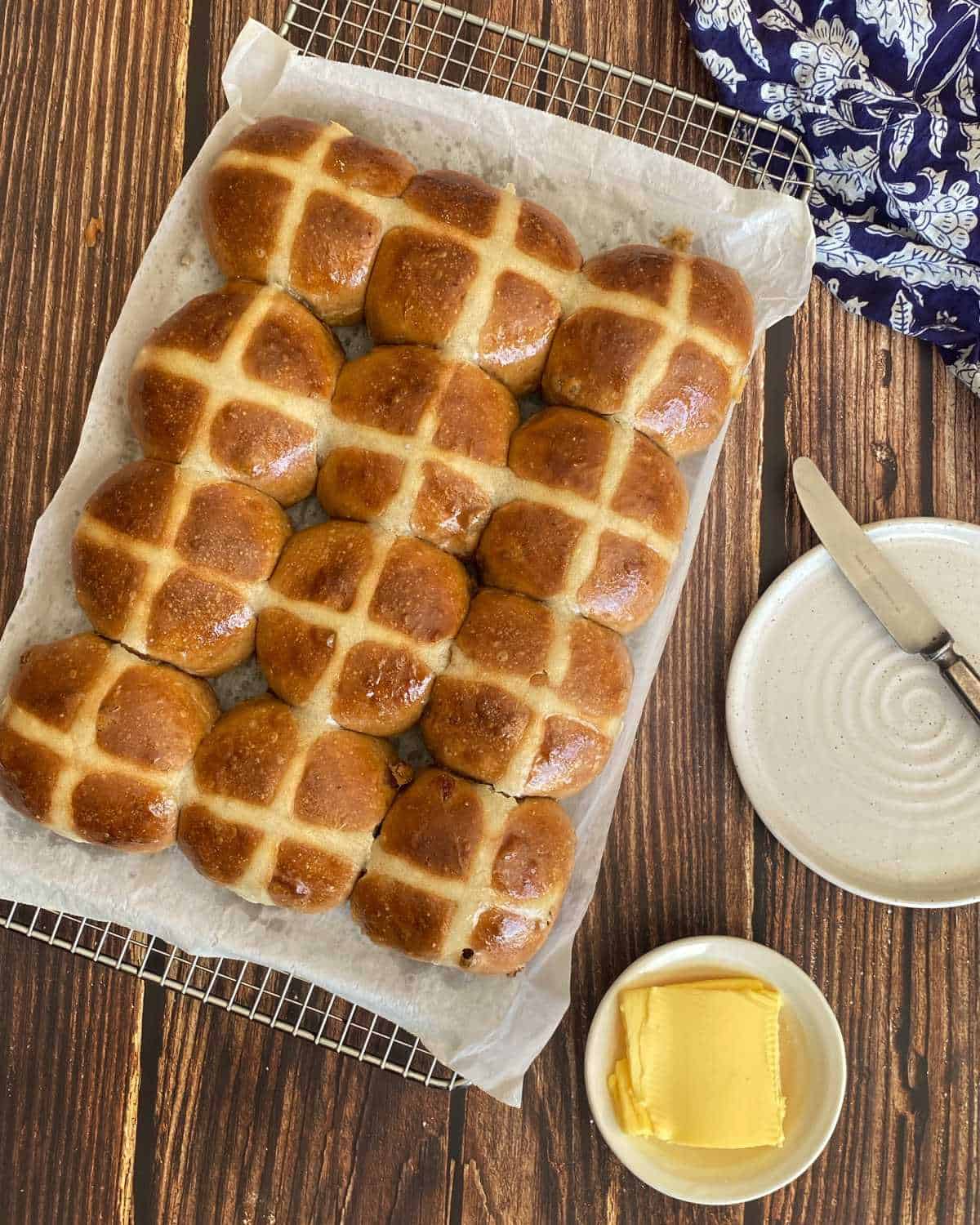 12 cooked hot cross buns sitting on a wire rack cooling. A small white plate with a knife lying across it and a small dish of butter to the side.