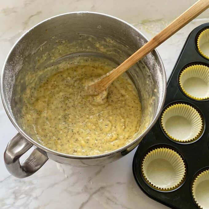 Lemon poppyseed mixture in a cake mixing bowl. A lined muffin tray with yellow muffin cases to the side.