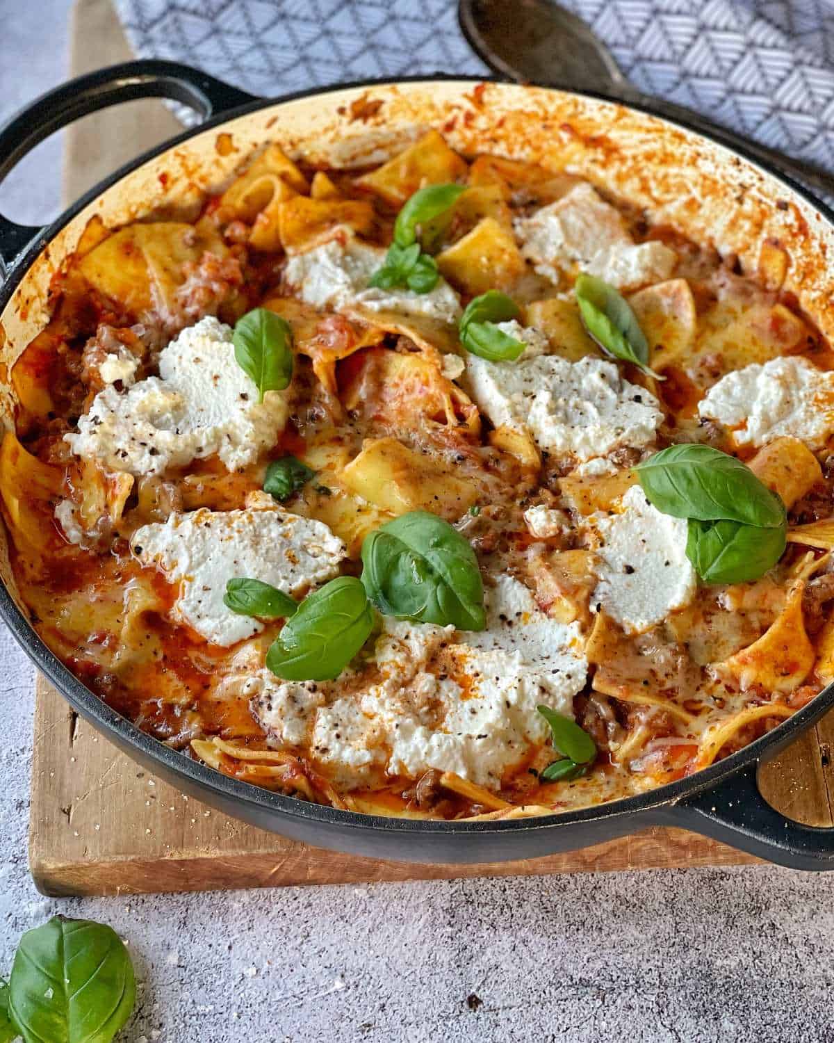 Cooked one pot lasagne in the round ceramic dish it was made in with scattered basil leaves and salt and pepper over the top sitting on a wooden chopping board.