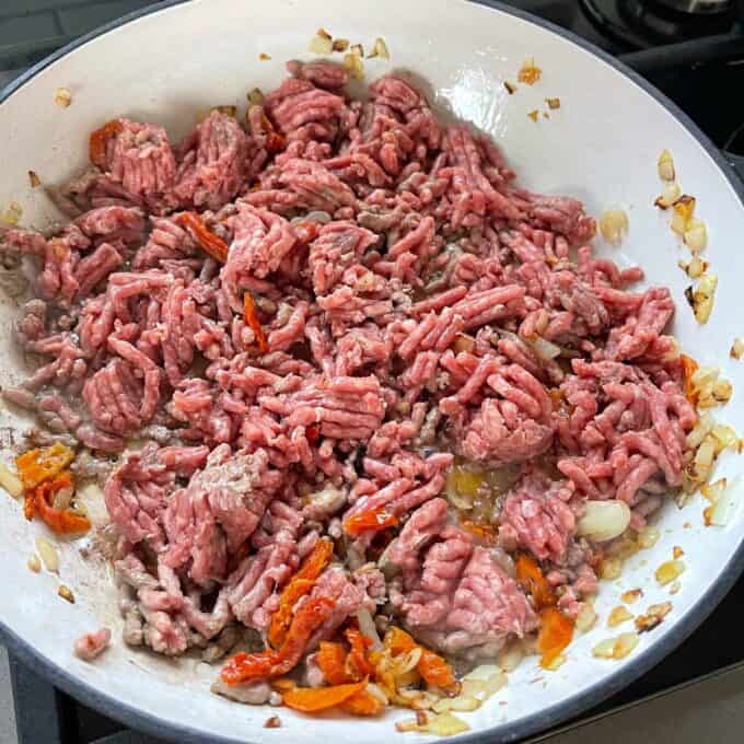 Mince, diced onion, crushed garlic, herbs and sundried tomatos frying in a round ceramic dish.