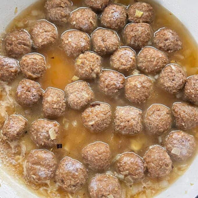 Meatballs, chopped onion and beef stock simmering in a round ceramic dish.