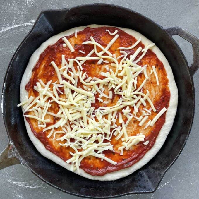 One uncooked pizza in a skillet frypan with pizza base and grated cheese on top.