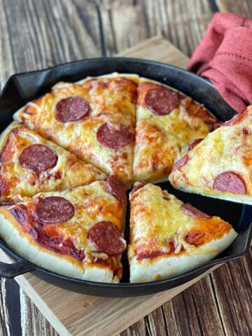 A cooked, sliced deep skillet pepperoni pizza in a skillet pan sitting on a wooden shopping board.