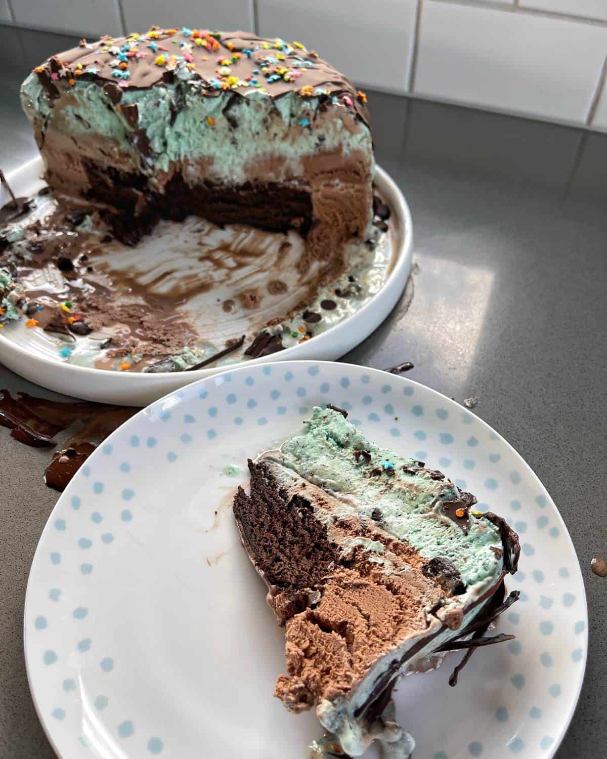 A piece of ice cream cake on a plate next to the remainder of the cake on another plate.