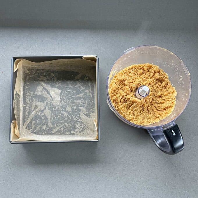 A lined baking tin and a food processor with a biscuit base on a grey bench.