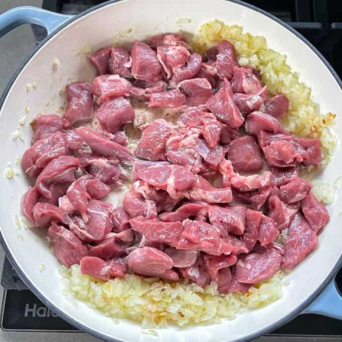Diced lamb being added to a round ceramic dish with fried onion and garlic.