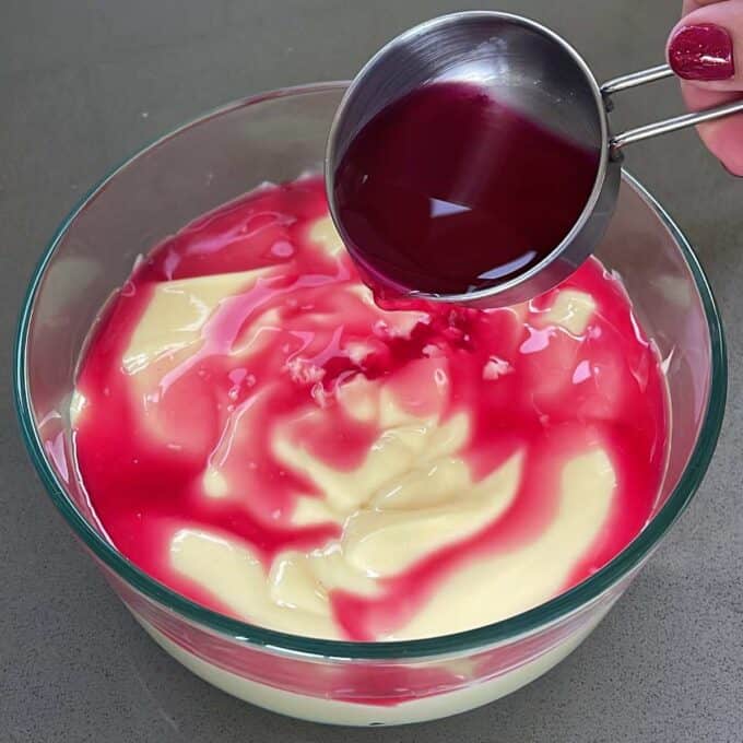 A glass bowl with custard - strawberry jelly is being poured over the top.