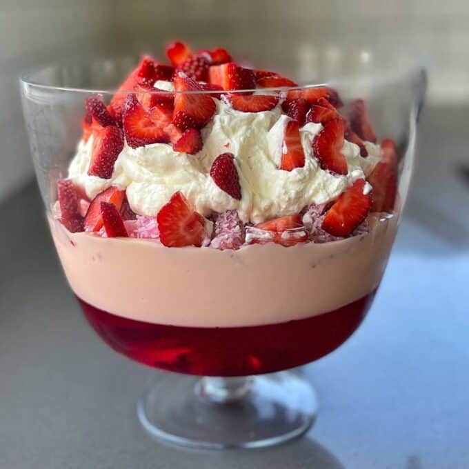 A close up shot of Strawbeery Lamington Triffle showing the different layers - strawberrry jelly, strawberry custard, sliced strawberry lamingtons, whipped cream and then topped with fresh sliced strawberries.