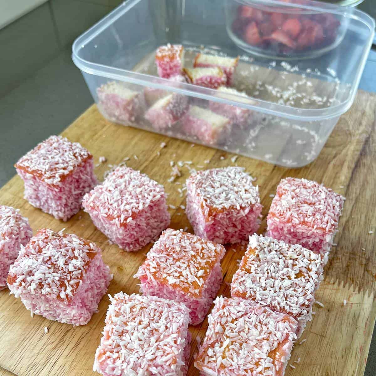 Strawberry lamingtons on a wooden chopping board. Slices lamingtons in a plastic container to the side.