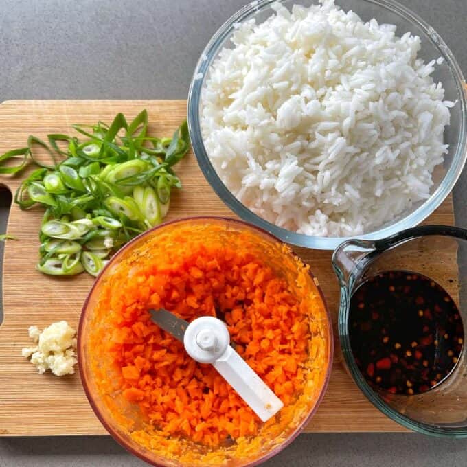 The chopped carrot, garlic and spring onion for Nasi Goreng sitting on a chopping board. A bowl of cooked whte rice amd sweet chilli sause in a small glass jug.