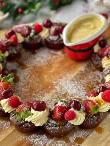 A close up of a brownie wreath served with cream and custard on a wooden chopping board.