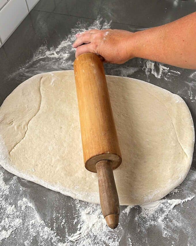 Pizza dough being rolled out on a floured bench with a rolling pin.