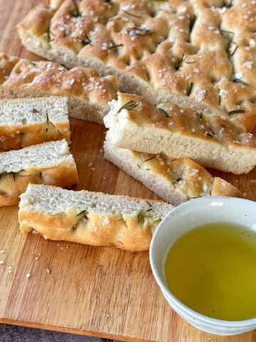 Focaccia bread thinly sliced and served on a woodedn chopping board with a sprig of rosemarry and a small dipping bowl of olive oil.