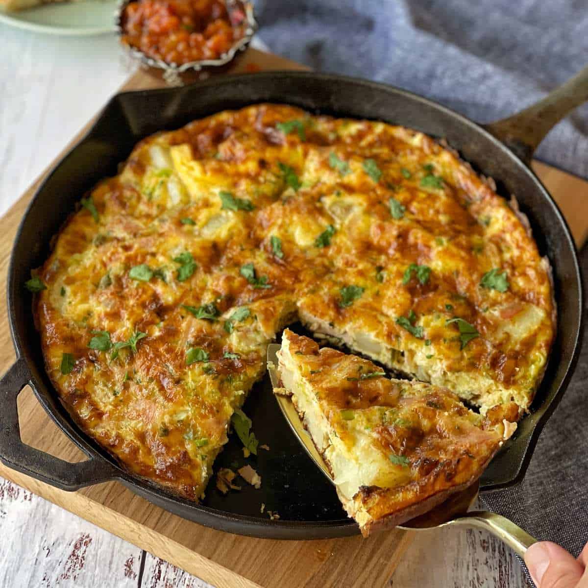 Ham and Potato Fritata in a round deep skillet pan with one slice removedand another silice being removed with a cake knife.