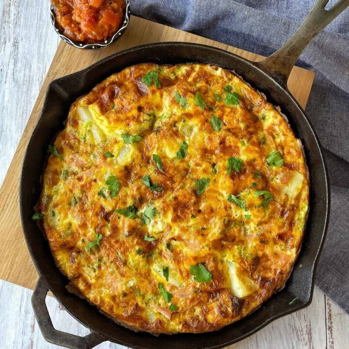 Ham and Potato Fritata in a round deep skillet pan on a wooden chopping board. A small dish of chutney served beside to accompany the fritata.
