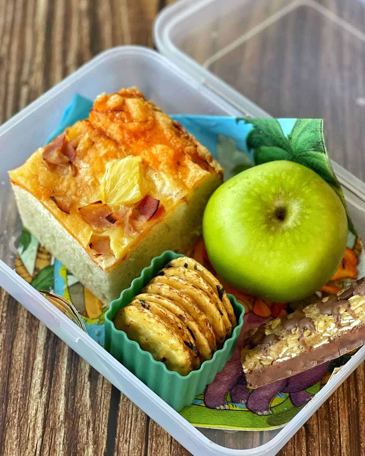 A slice of pizza bread in a lunch box with an apple and some rice crackers.