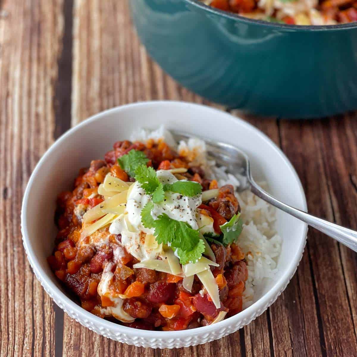 A serving of Vegetarian Chilli in a white bowl served with rice and a dash of sour cream, gratted cheese and corainder leaves.