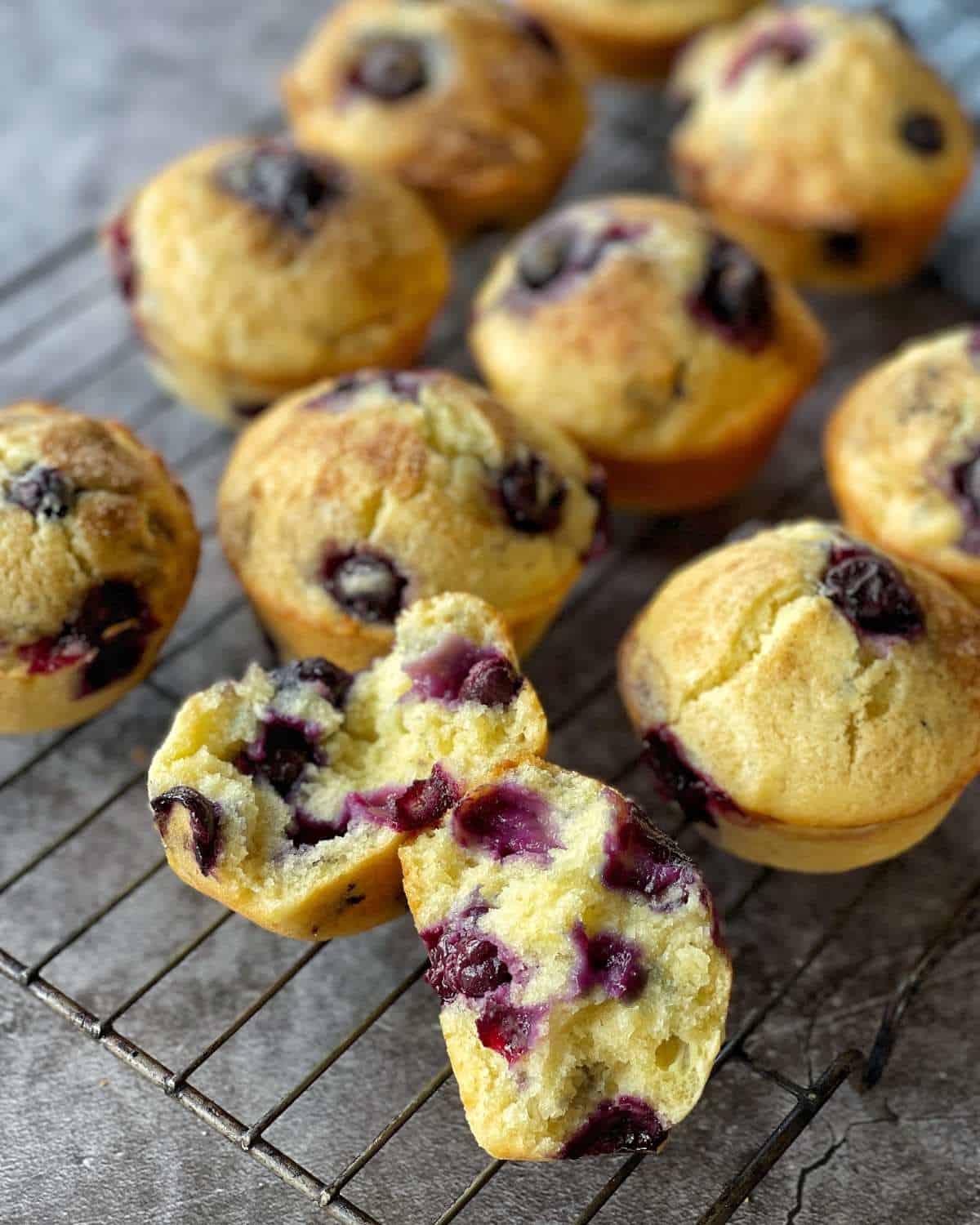 A selection of cooked blueberry muffins on a cooling rack. One muffin has been split open to show the blueberry filling.