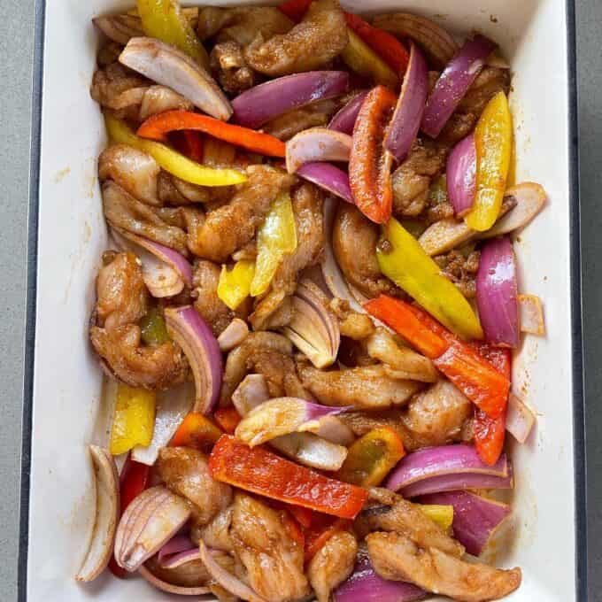 Uncooked marinated vetegtables and chicken peices to make Chicken Fajita Tray Bake in a over proof dish sitiing on a grey bench top.