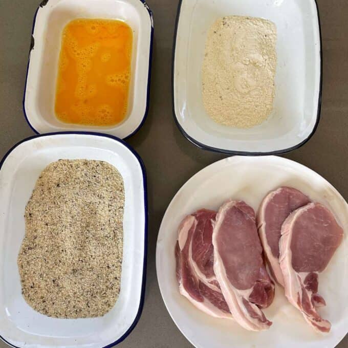 Four dishes sitting on a grey bench top- one with flour, one with egg, one with breadcrumbs and one with the pork chops.