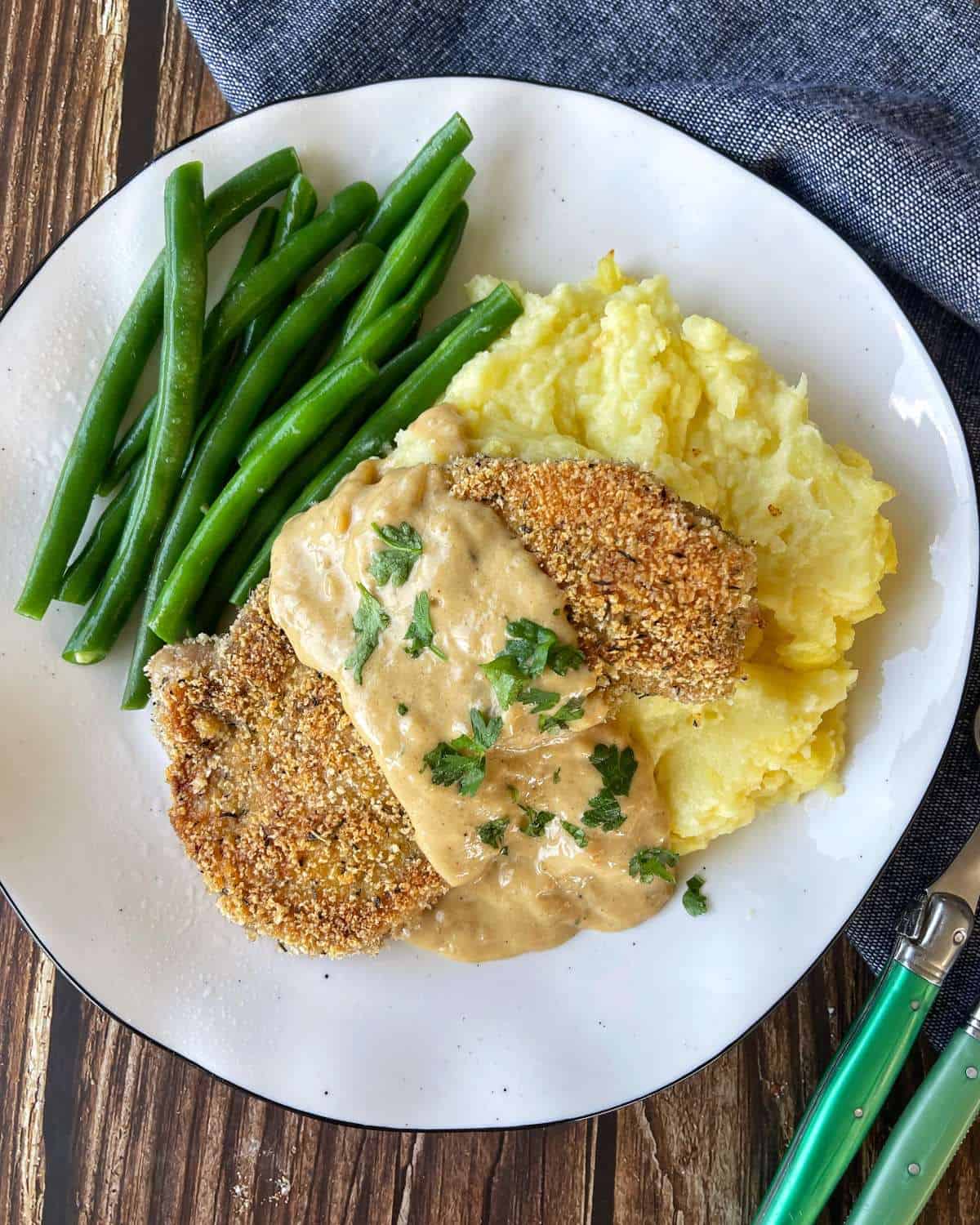 One cooked crumbed pork chop served with mashed potato and green beans. A creamy gravey has been poured over the top as well and some parsley leaves.