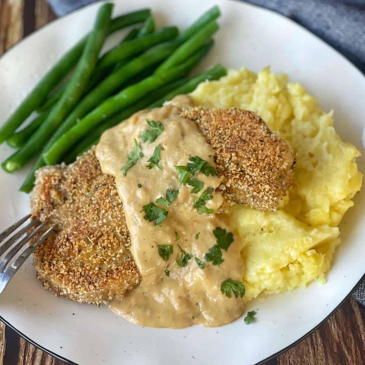 One cooked crumbed pork chop served with mashed potato and green beans. A creamy gravey has been poured over the top as well and some parsley leaves.