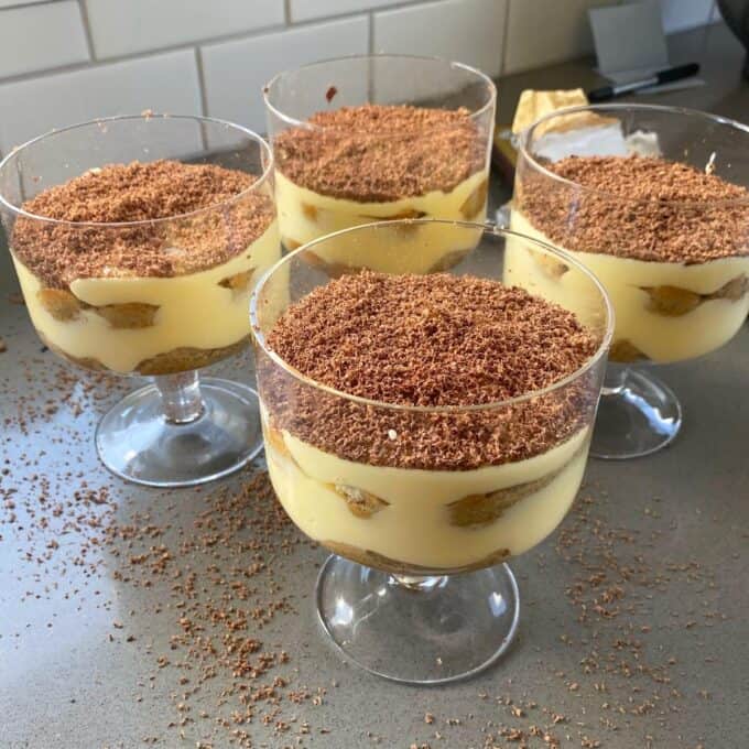 Four individual tiramisu's served in small glasses sitting on a grey bench. Grated chocolate has just been layered over the top.