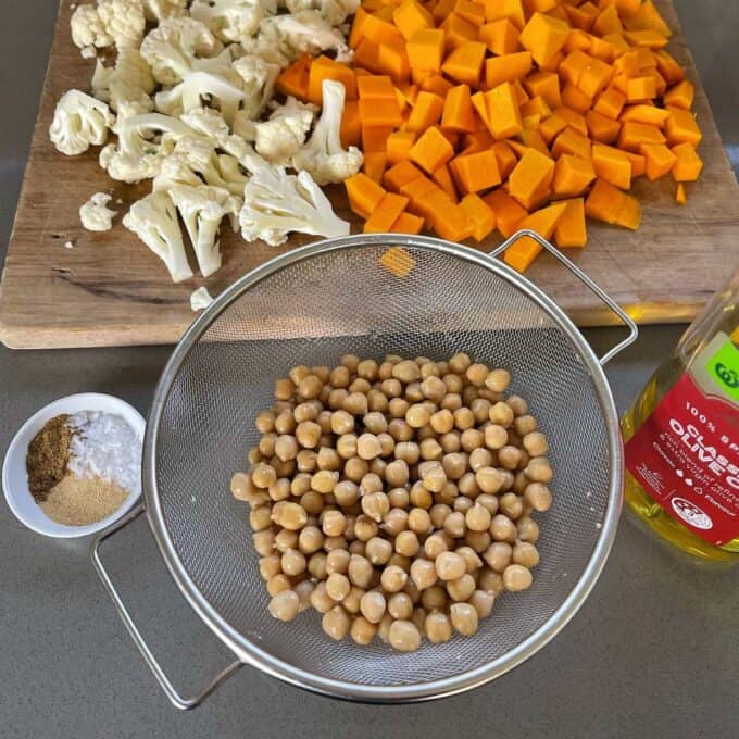 Cubed peices of pumpkin and small peices of cauliflower on a wooden chopping board. Strained and rinced chickpeas in a sieve and a small white bowl of salt and spices all sitting on a grey bench top to make Nourish Bowls.