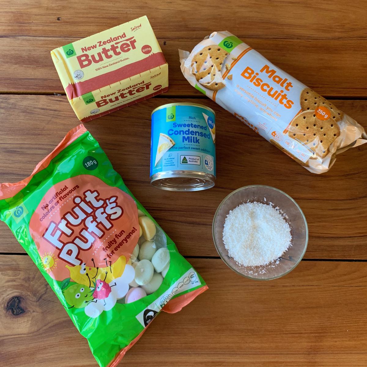 Butter, malt biscuits, a can of sweetened condensed milk, a bag of fruit puffs, and a bowl of desiccated coconut—all the classic ingredients for making delicious Lolly cake—arranged on a wooden surface.
