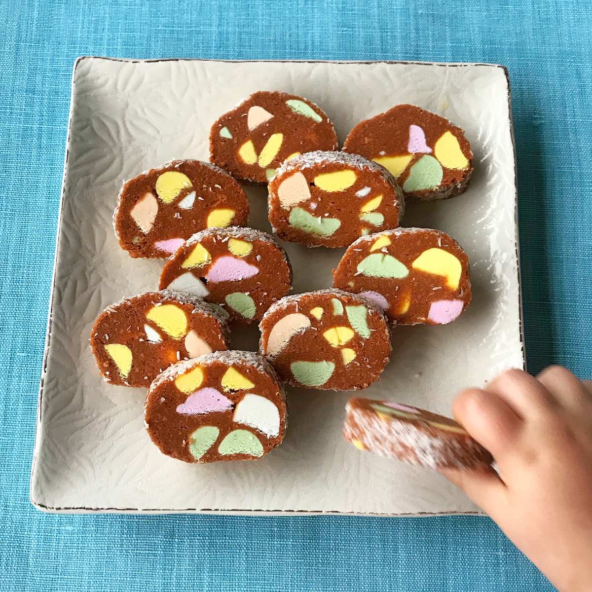 A square plate with nine slices of chocolate marshmallow dessert, similar to lolly cake, sits on a blue surface; a child’s hand is reaching for one slice.
