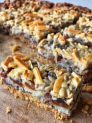 Close-up of magic slice dessert bars with a crumbly base, visible layers of dates, and a topping of sliced almonds and coconut on a wooden surface.