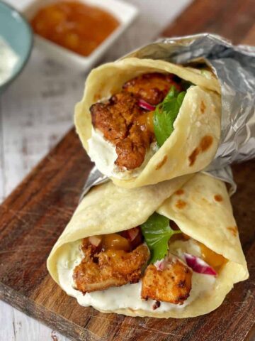 Two cooked air fryer tandoori chicken flatbreads wrapped in tinfoil and served on a wooden chopping board