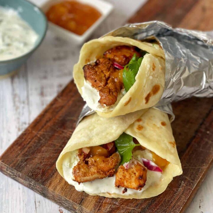 Two cooked air fryer tandoori chicken flatbreads wrapped in tinfoil and served on a wooden chopping board