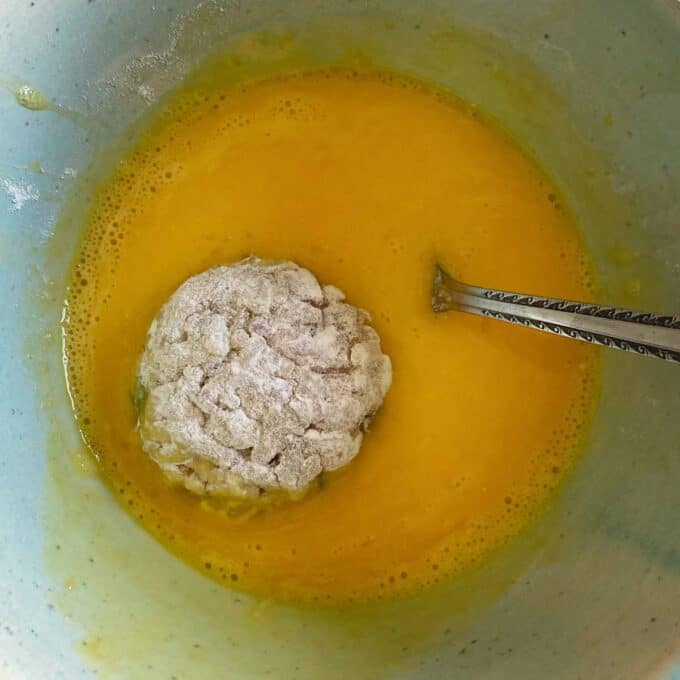 A rolled and floured arancini being dipped into a mixed egg