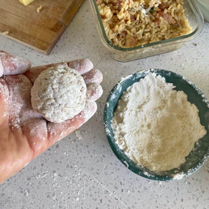 A hand rolling an arancini ball through flour