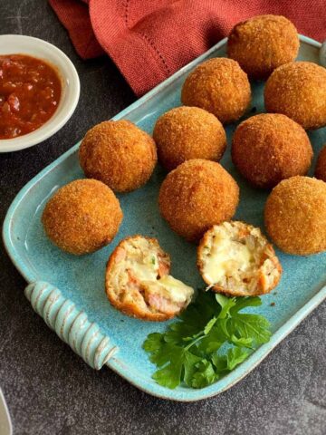 A selection of cooked arancini balls on a duck egg blue platter served with parsley. One Arancini balls has been split open showing the gooey mozzarella filling