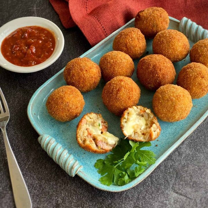 A selection of cooked arancini balls on a duck egg blue platter served with parsley. One Arancini balls has been split open showing the gooey mozzarella filling
