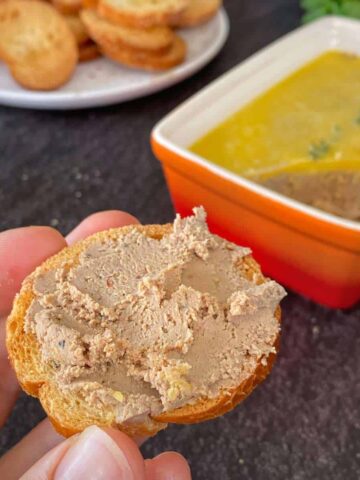 A close up shot of a hand holding a piece of crostini with chicken liver spread over the top
