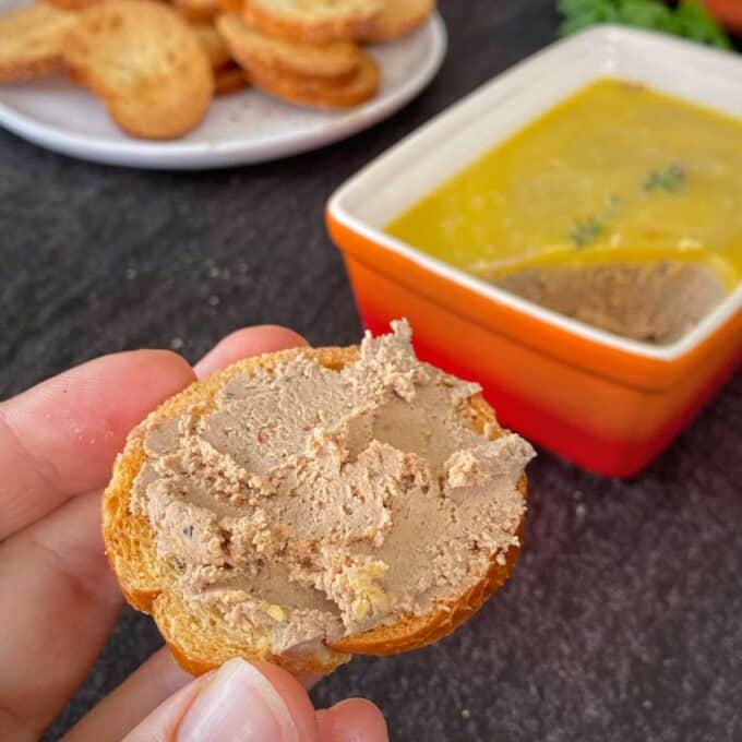A close up shot of a hand holding a piece of crostini with chicken liver spread over the top