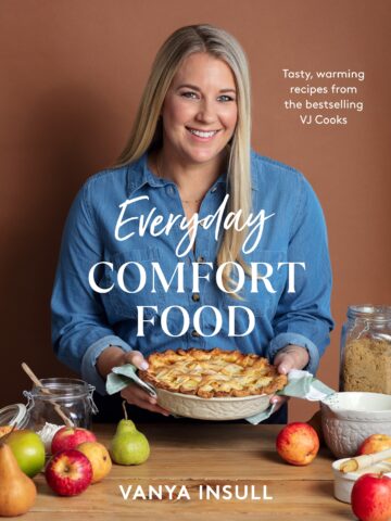 A blonde woman standing in her kitchen holding a apple pie. There are apples and baking equipment on the table in front of her.