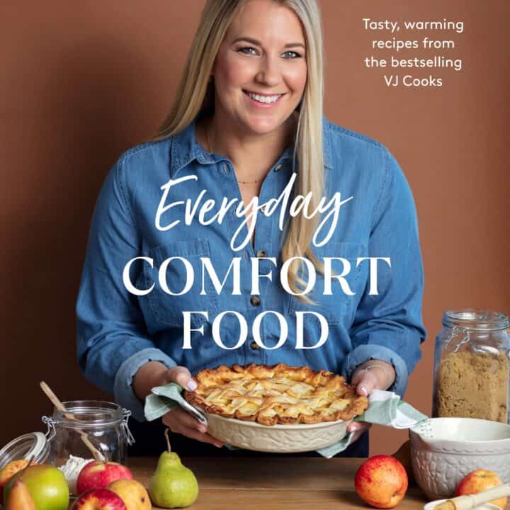 A blonde woman standing in her kitchen holding a apple pie. There are apples and baking equipment on the table in front of her.