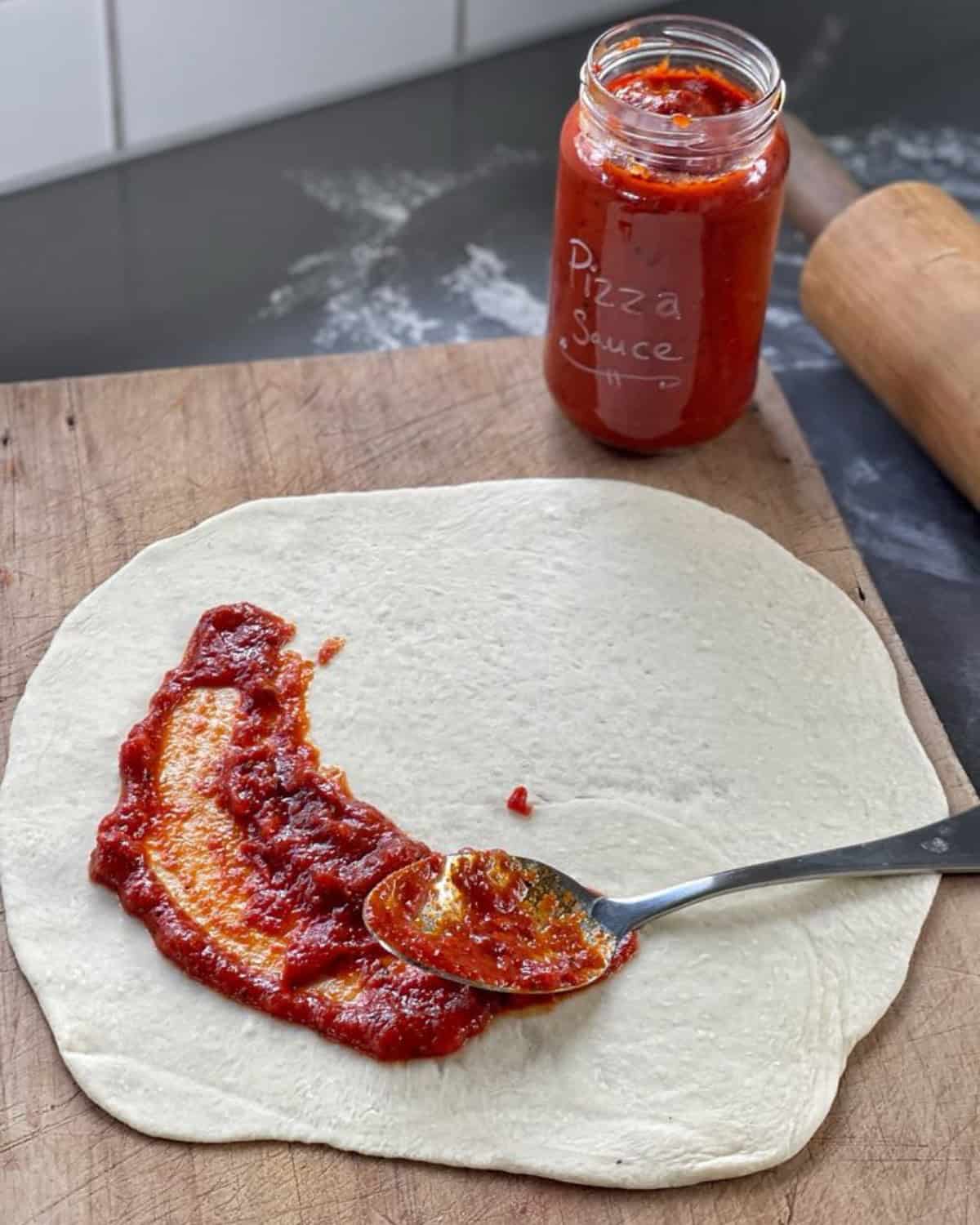 Pizza sauce being spread on a pizza base on a wooden chopping board.