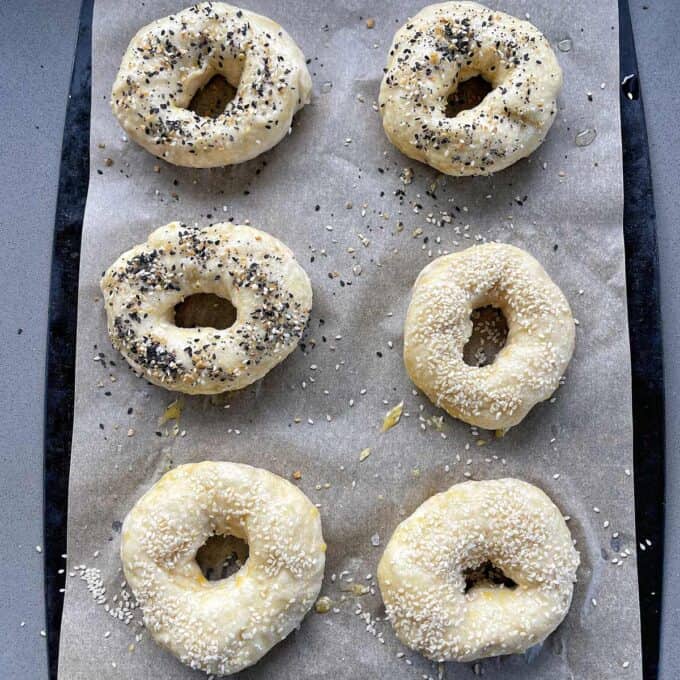 Six uncooked bagels on parchment paper, topped with sesame and mixed seeds, on a baking tray.