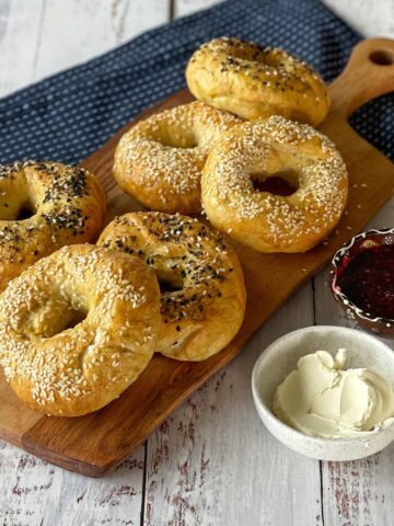 Bagels with sesame and poppy seeds on a wooden board, accompanied by cream cheese and a small bowl of red jam on a rustic white table with a blue cloth napkin.