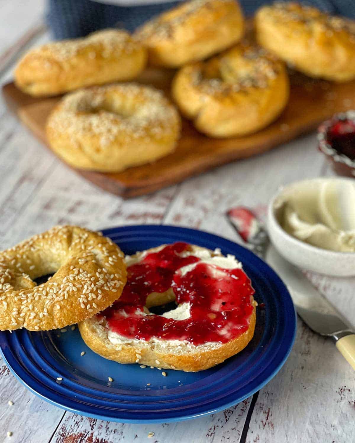 A bagel with cream cheese and red jam on a blue plate. A knife and a bowl of cream cheese are beside it. More bagels are visible on a wooden board in the background.